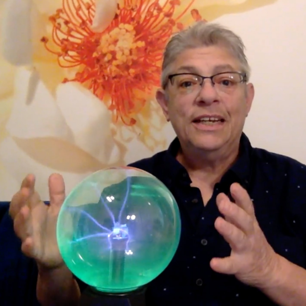 Miche wearing a black shirt wearing glasses with hands hovering over plasma ball with electrical discharges touching glass from center of the ball.
